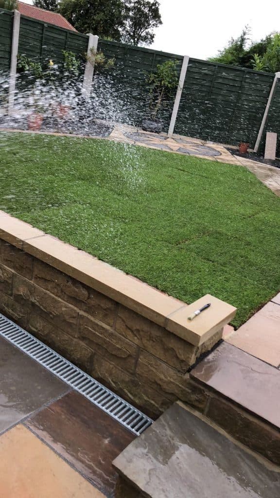 A sprinkler watering a newly laid lawn above a stone retaining wall, with a drainage channel set into the paving below