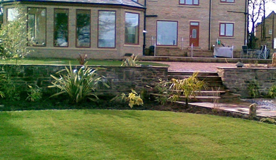 A stone retaining wall terracing a garden below a house, with wide stone steps up to the paved area and a level lawn below