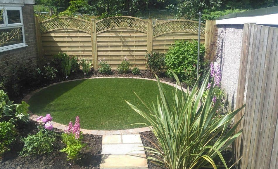 An oval lawn edged with a curved sandstone path, surrounded by newly planted borders under a trellis-topped fence