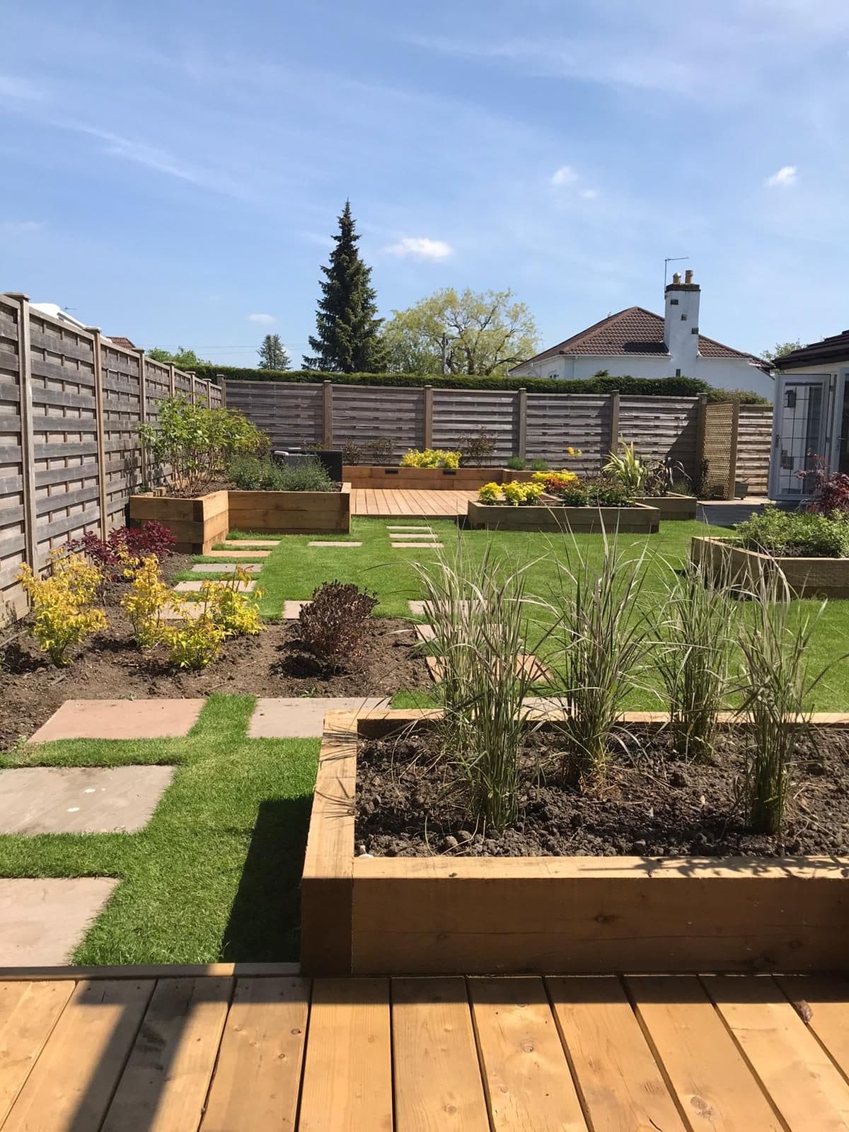 Timber sleeper raised beds filled with fresh compost beside a lawn, stepping-stone paving and a deck